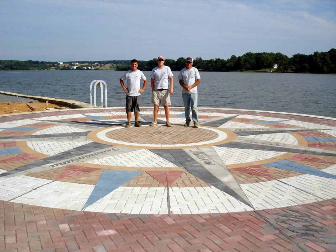 Three Stepping Stone Hardscape crew members standing on the finished compass rose paver plaza at Leonardtown Wharf, with the river behind them